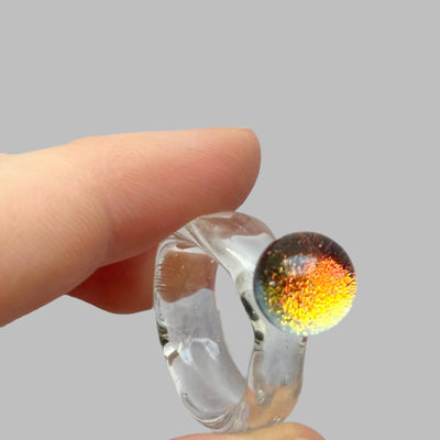 Clear glass ring with colorful interior held between fingers against a gray background