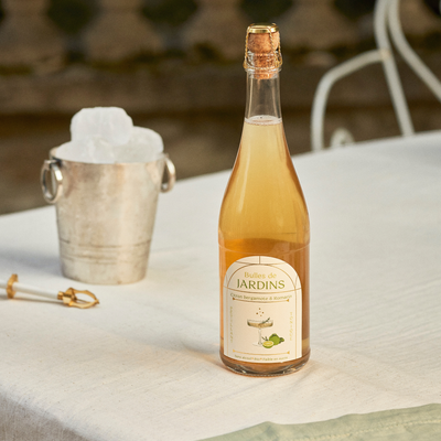 Bottle of Bulles de Jardins alcohol-free sparkling wine with bergamot and rosemary, next to an ice bucket and champagne opener on a white tablecloth.