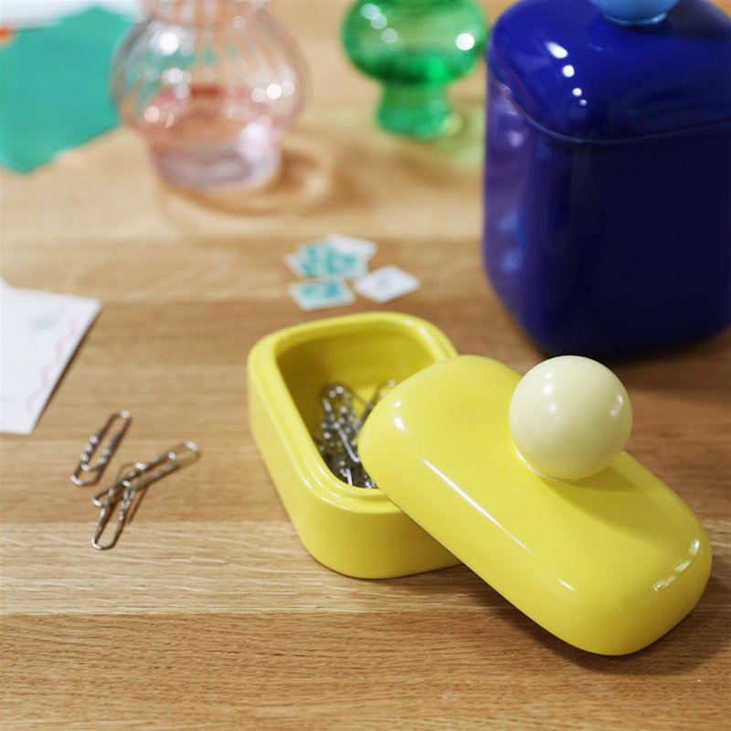 a small yellow storage jar with a spherical handle on top, holding paper clips, sits on top of a wooden desk