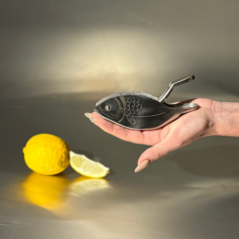 Hand holding a metal fish-shaped lemon squeezer with a lemon and lime on a reflective surface.