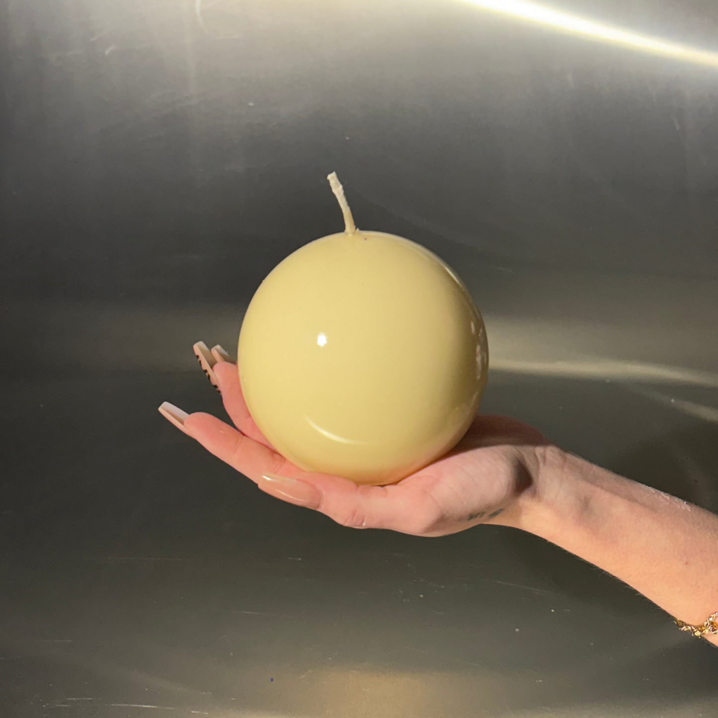 Hand holding a spherical yellow candle against a reflective metallic background