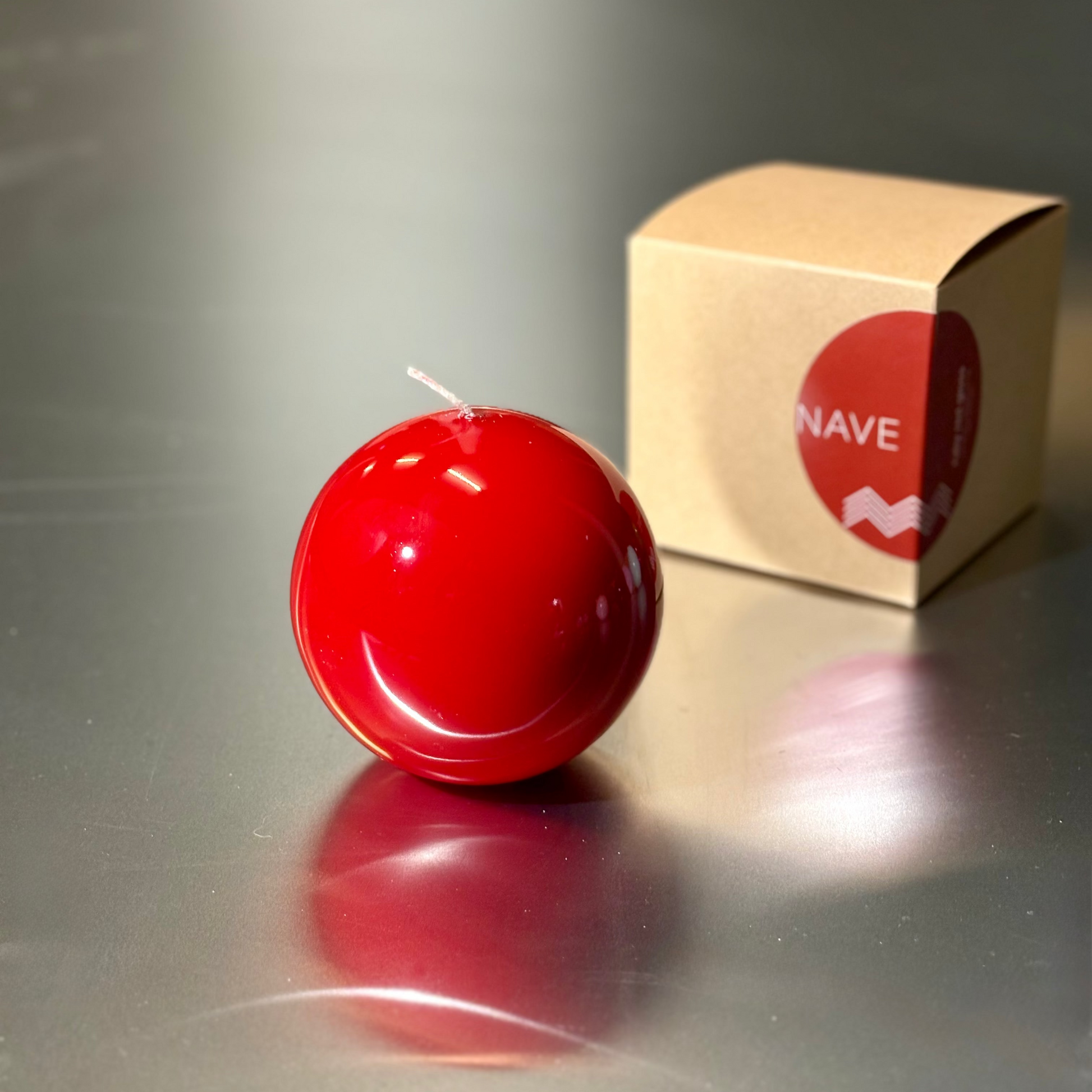 Red spherical candle on a reflective surface with a beige box labeled 'Nave' in the background.
