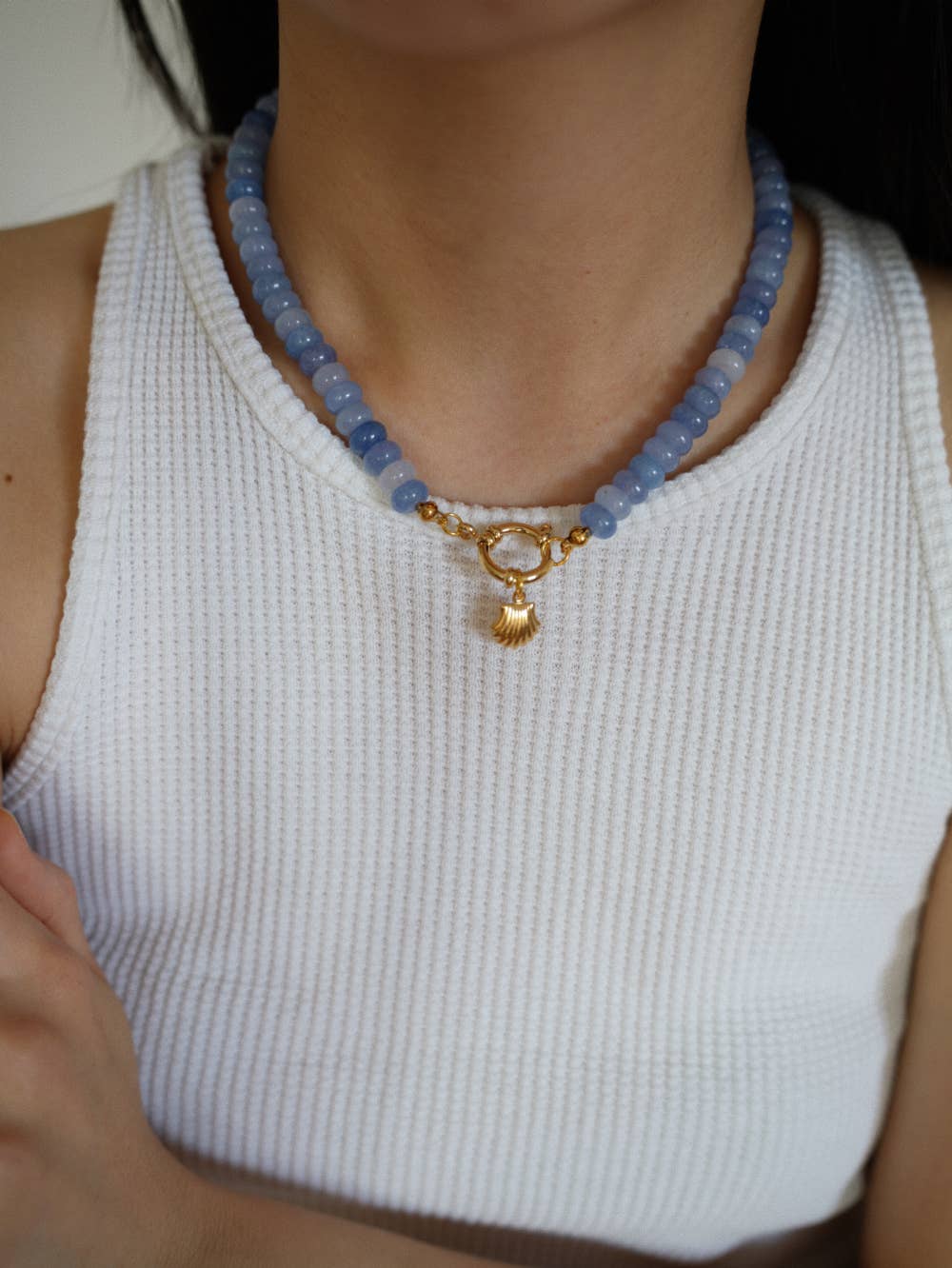 Person wearing a blue beaded necklace with a gold clasp on a white sleeveless top.