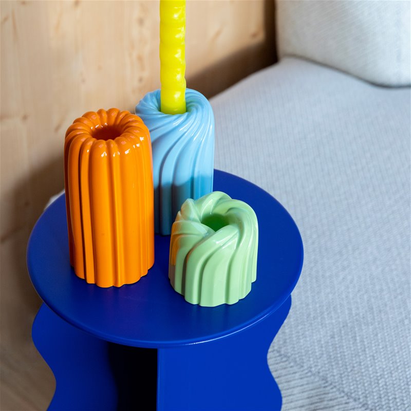 three brightly coloured candle holders with a swirly pattern inspired by bundt cake, each in a different size in light blue, orange and green on a trendy blue coffee table