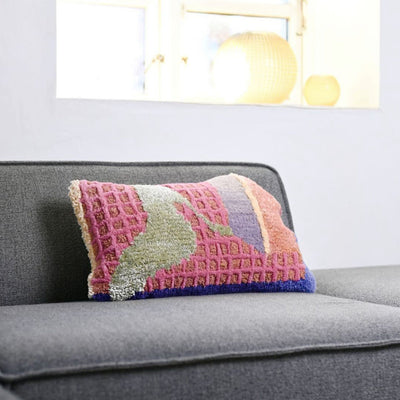 waffle cushion has a squashed rectangular shape with an organic shaped edge, the pattern is abstract with a waffle like 3d structure and pastel shades of green, lilac and pink on a grey couch